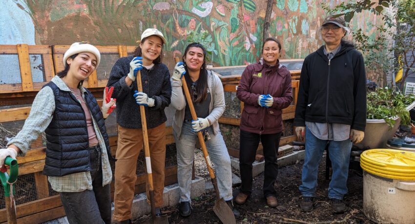 Five people stand in a garden smiling at the camera. Three of them are holding shovels. Behind them is a large wooden composting bin and a wall mural.
