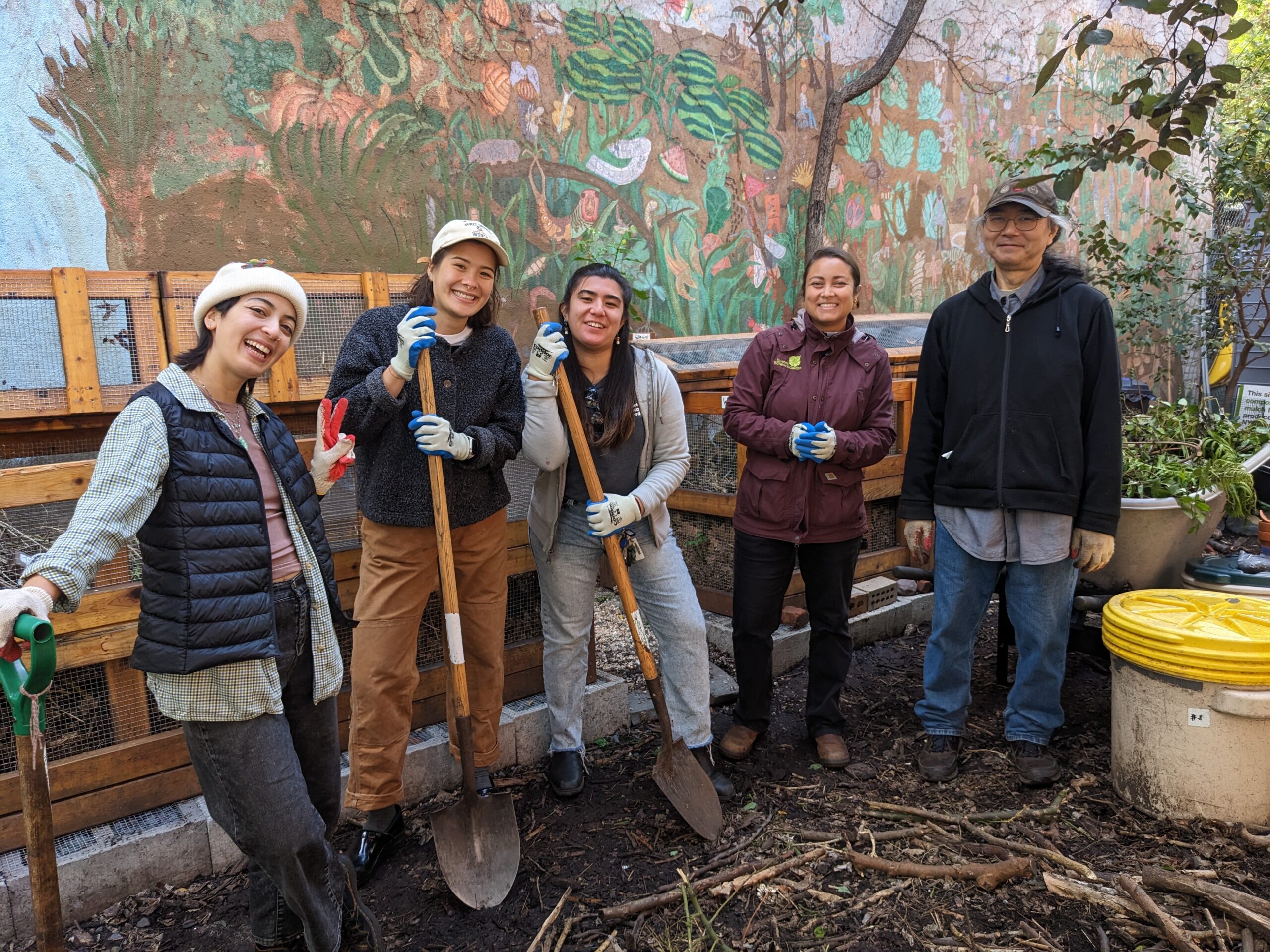 Five people stand in a garden smiling at the camera. Three of them are holding shovels. Behind them is a large wooden composting bin and a wall mural.