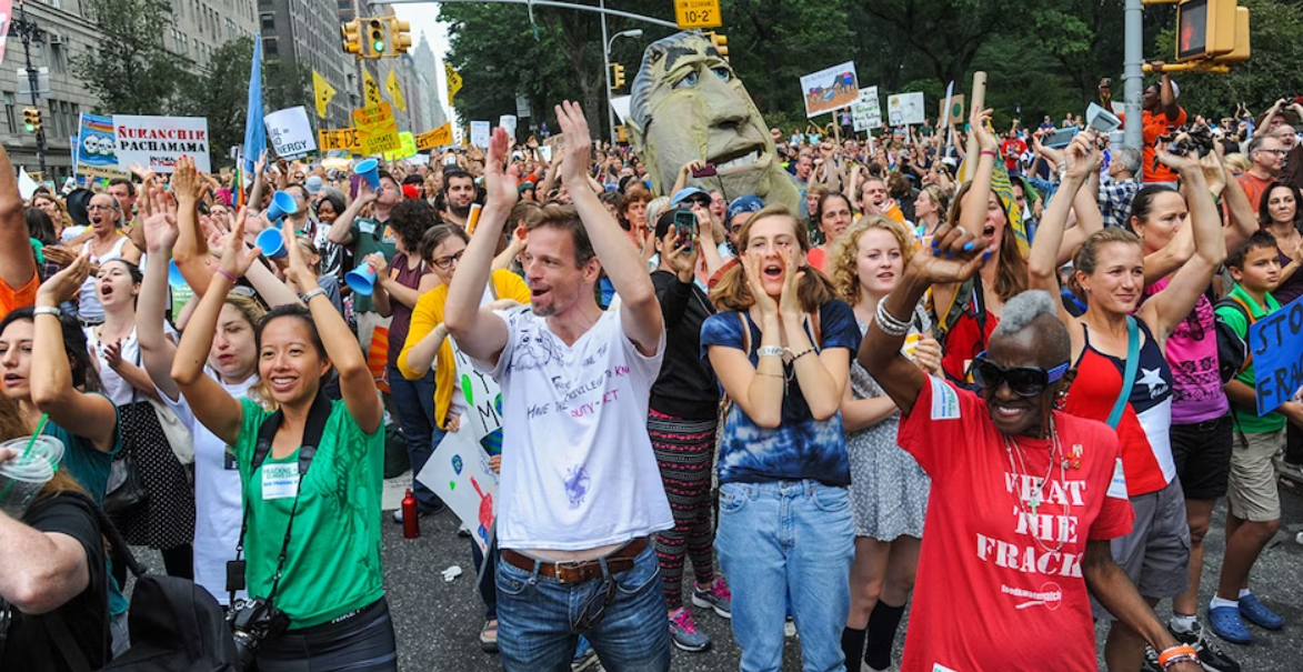 A large crowd of people marching down an avenue in New York holding banners protesting the continued use of fossil fuels