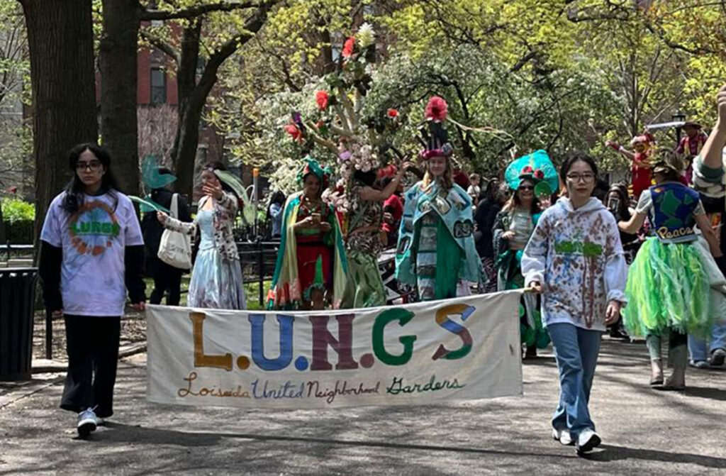 Image of the head of a LUNGS Spring Awakening Parade in Tompkins Square Park with a large banner and people dressed in Earth Day-themed costumes.